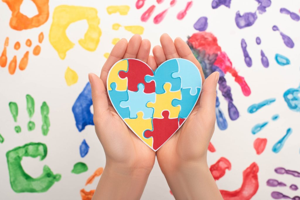 cropped view of woman holding heart with puzzle on white with hand prints for world autism awareness - Channel Islands Family Dental Office | Dentist In Ventura County